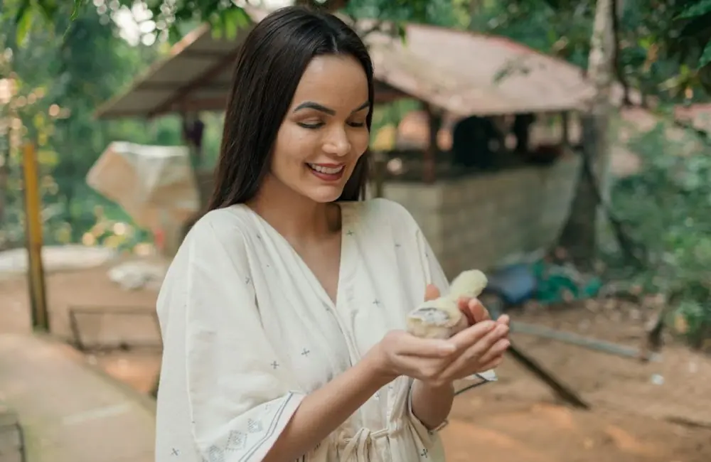 Smiling woman in a light kurta gently holding a baby chick at a lush Kerala Ayurveda retreat garden with rustic cottages in the background.