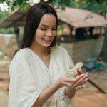 Smiling woman in a light kurta gently holding a baby chick at a lush Kerala Ayurveda retreat garden with rustic cottages in the background.