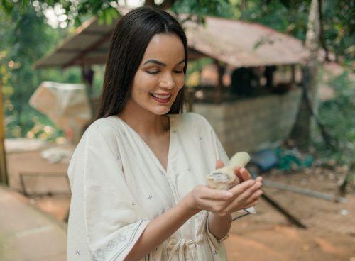 Smiling woman in a light kurta gently holding a baby chick at a lush Kerala Ayurveda retreat garden with rustic cottages in the background.