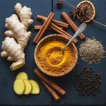 Overhead flat lay of winter spices—fresh ginger root and slices, a wooden bowl of turmeric powder with a spoon, cinnamon sticks, cumin seeds, black peppercorns, star anise, and a dried orange slice—arranged on a dark slate surface.