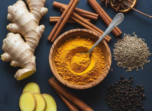 Overhead flat lay of winter spices—fresh ginger root and slices, a wooden bowl of turmeric powder with a spoon, cinnamon sticks, cumin seeds, black peppercorns, star anise, and a dried orange slice—arranged on a dark slate surface.