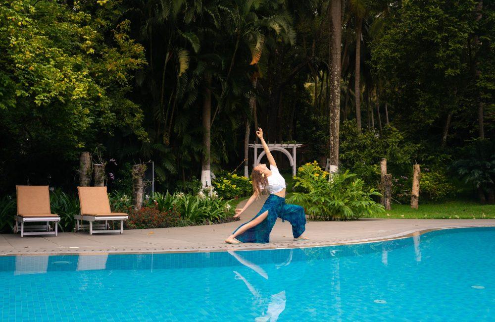 Woman practicing a yoga backbend pose beside a blue swimming pool, surrounded by lush tropical greenery in Kerala.