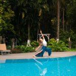 Woman practicing a yoga backbend pose beside a blue swimming pool, surrounded by lush tropical greenery in Kerala.