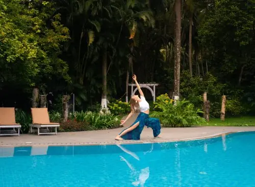 Woman practicing a yoga backbend pose beside a blue swimming pool, surrounded by lush tropical greenery in Kerala.