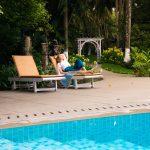Guest relaxing on a sun lounger beside a blue-tiled pool surrounded by lush tropical greenery.