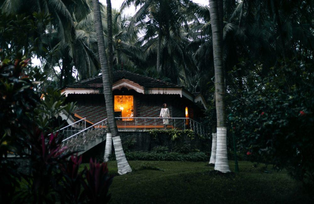 A traditional Kerala-style cottage surrounded by tall coconut palms at dusk, with warm light glowing from a window and a guest standing on the veranda.