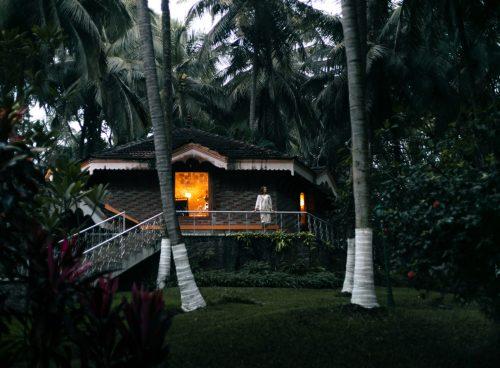 A traditional Kerala-style cottage surrounded by tall coconut palms at dusk, with warm light glowing from a window and a guest standing on the veranda.