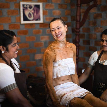 Smiling guest after a herbal body therapy sits with two Ayurvedic therapists at Kairali – The Ayurvedic Healing Village.