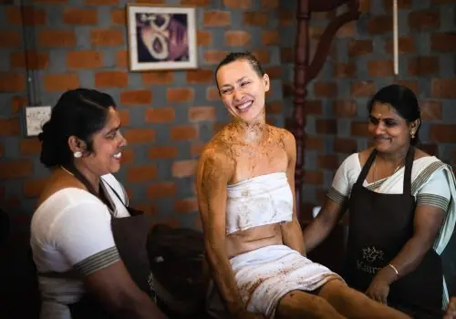 Smiling guest after a herbal body therapy sits with two Ayurvedic therapists at Kairali – The Ayurvedic Healing Village.