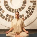 A person sits cross-legged in meditation pose with eyes closed, wearing a cream kurta and pants, against a circular stone-patterned wall.