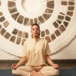A person sits cross-legged in meditation pose with eyes closed, wearing a cream kurta and pants, against a circular stone-patterned wall.