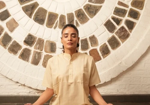A person sits cross-legged in meditation pose with eyes closed, wearing a cream kurta and pants, against a circular stone-patterned wall.