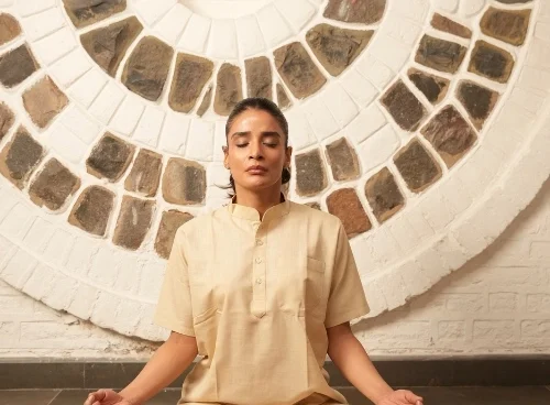 A person sits cross-legged in meditation pose with eyes closed, wearing a cream kurta and pants, against a circular stone-patterned wall.