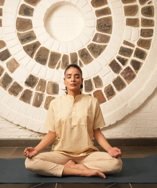 A person sits cross-legged in meditation pose with eyes closed, wearing a cream kurta and pants, against a circular stone-patterned wall.