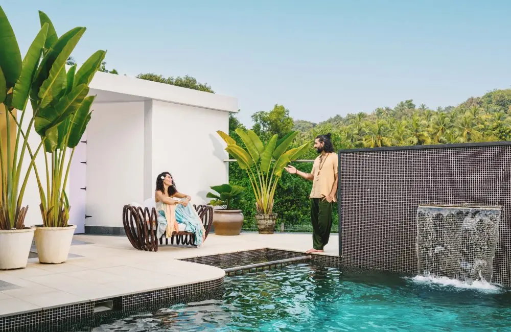 Couple relaxing by the infinity pool at Villa Raag in South Goa, surrounded by tropical greenery and a waterfall feature.