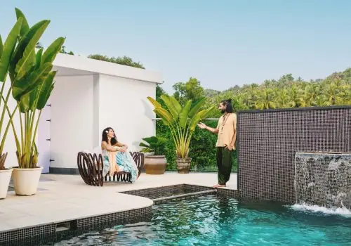 Couple relaxing by the infinity pool at Villa Raag in South Goa, surrounded by tropical greenery and a waterfall feature.