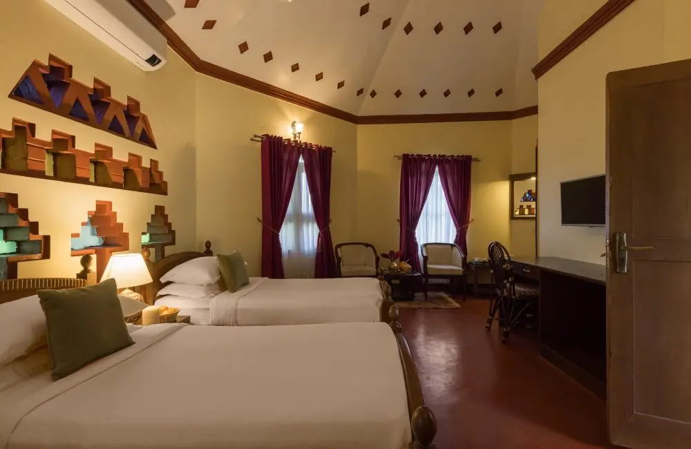 Twin-bed guest room at Kairali – The Ayurvedic Healing Village in Palakkad, featuring warm lighting, traditional Kerala-inspired wall niches, high vaulted ceiling, seating area, and maroon curtains.