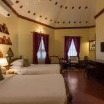 Twin-bed guest room at Kairali – The Ayurvedic Healing Village in Palakkad, featuring warm lighting, traditional Kerala-inspired wall niches, high vaulted ceiling, seating area, and maroon curtains.