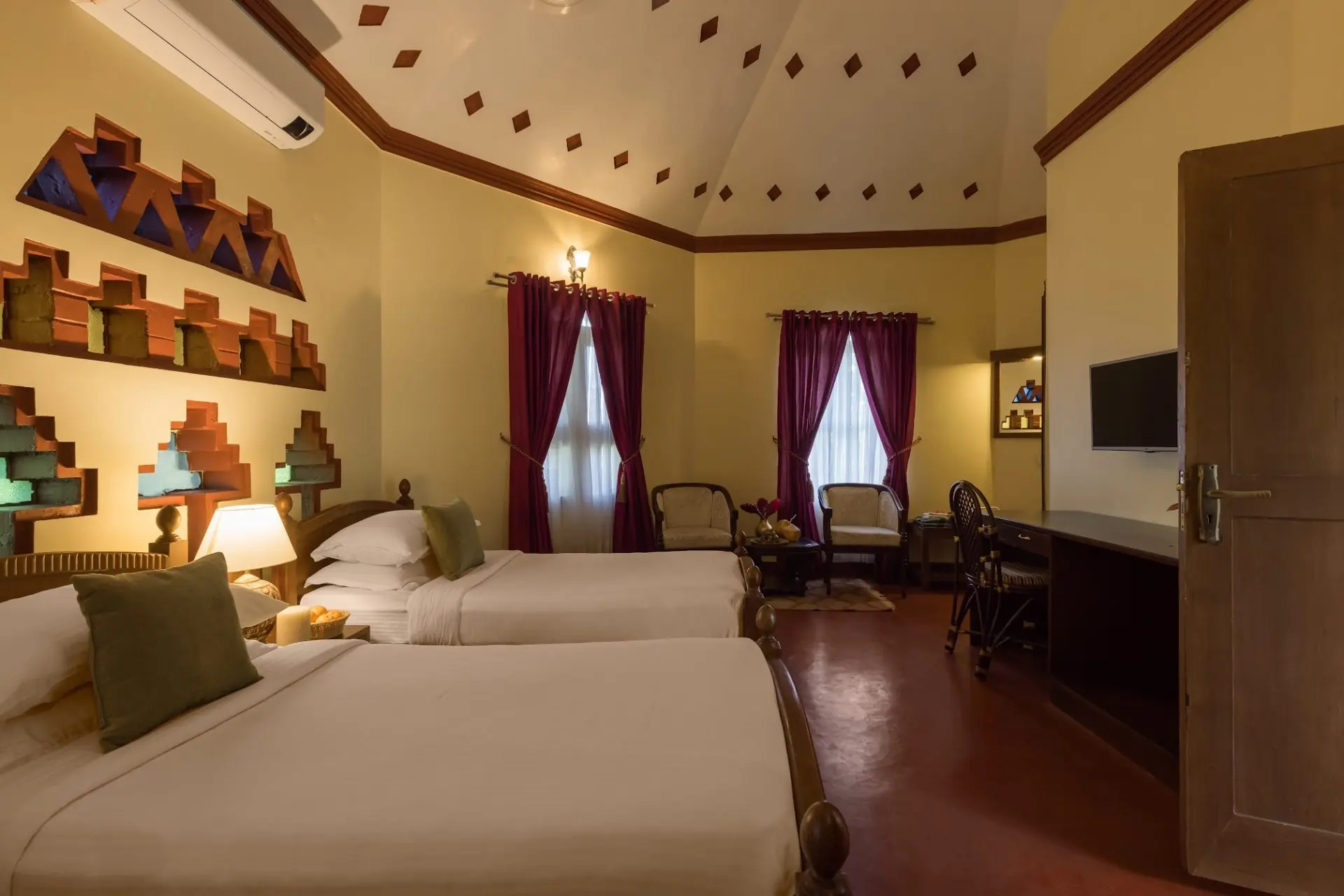 Twin-bed guest room at Kairali – The Ayurvedic Healing Village in Palakkad, featuring warm lighting, traditional Kerala-inspired wall niches, high vaulted ceiling, seating area, and maroon curtains.