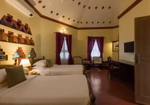 Twin-bed guest room at Kairali – The Ayurvedic Healing Village in Palakkad, featuring warm lighting, traditional Kerala-inspired wall niches, high vaulted ceiling, seating area, and maroon curtains.