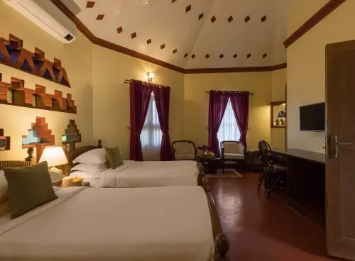 Twin-bed guest room at Kairali – The Ayurvedic Healing Village in Palakkad, featuring warm lighting, traditional Kerala-inspired wall niches, high vaulted ceiling, seating area, and maroon curtains.