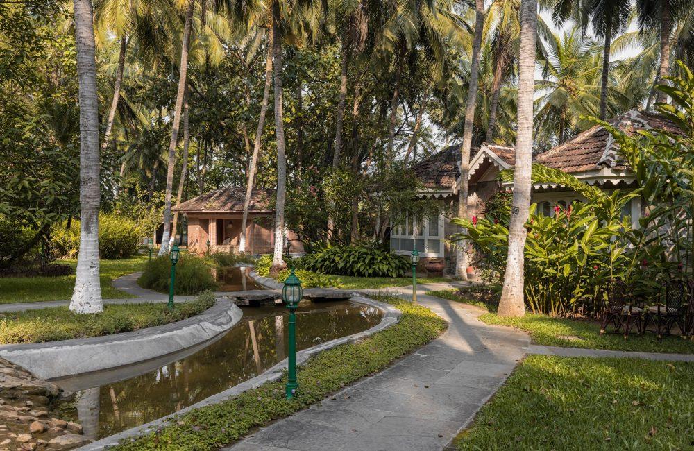 Palm-lined garden walkway at Kairali – The Ayurvedic Healing Village in Palakkad, with heritage cottages beside a serene water channel in a lush Kerala landscape.