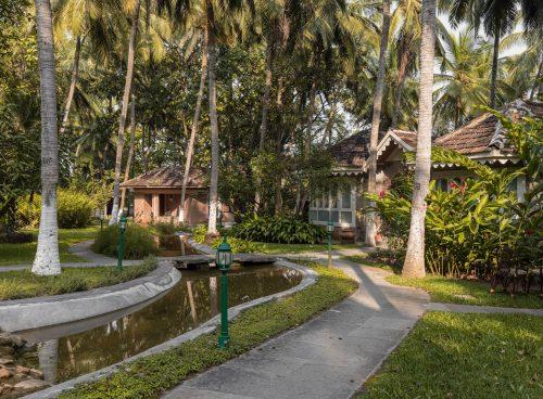Palm-lined garden walkway at Kairali – The Ayurvedic Healing Village in Palakkad, with heritage cottages beside a serene water channel in a lush Kerala landscape.