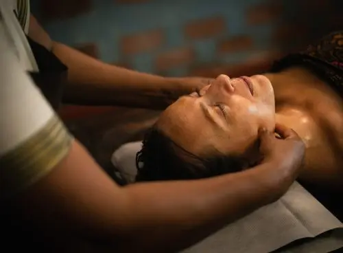 A woman receiving a traditional Ayurvedic head and neck massage at a spa, lying on a treatment table with eyes closed and turmeric oil applied to her face