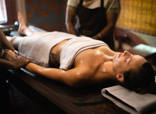 A woman lying on a wooden massage table receiving an Ayurvedic oil massage from two therapists in a traditional setting.
