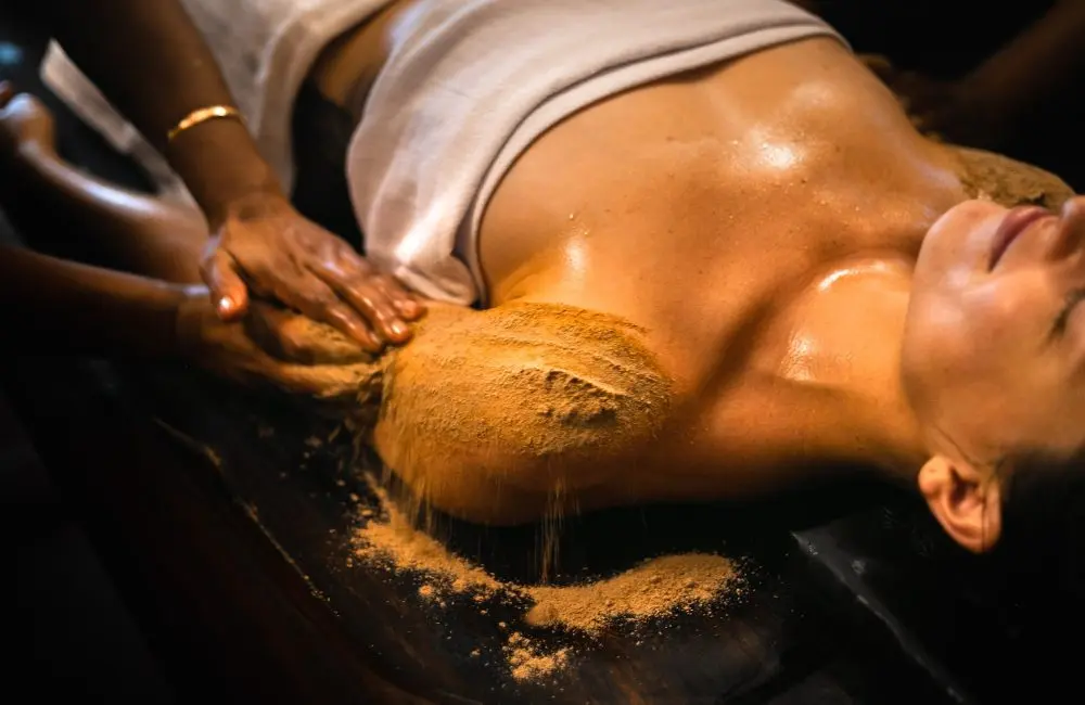Ayurvedic therapist performing an herbal powder massage on a woman lying on a wooden treatment table, with warm lighting highlighting the oiled skin and cascading powder on her arm.