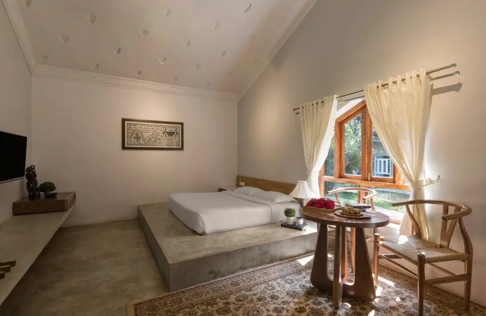 Sunlit Vaastu villa bedroom at Kairali Ayurvedic Healing Village featuring a low platform bed, wooden window, sheer curtains, and a small seating table with fruits.