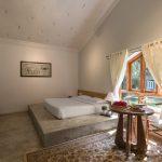 Sunlit Vaastu villa bedroom at Kairali Ayurvedic Healing Village featuring a low platform bed, wooden window, sheer curtains, and a small seating table with fruits.