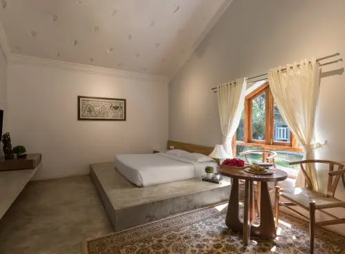 Sunlit Vaastu villa bedroom at Kairali Ayurvedic Healing Village featuring a low platform bed, wooden window, sheer curtains, and a small seating table with fruits.