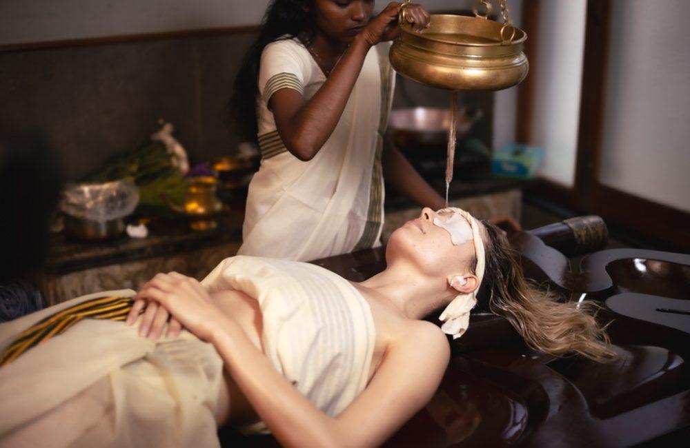 A female Ayurvedic therapist in a white sari pours warm herbal oil from a brass pot suspended by chains onto the forehead of a relaxed client lying on a wooden table, eyes covered with white cloth, in a serene treatment room with wooden elements and herbs nearby.
