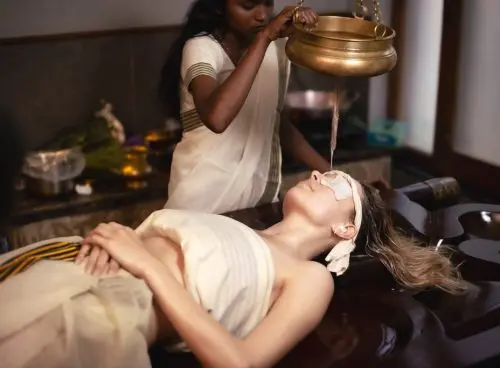 A female Ayurvedic therapist in a white sari pours warm herbal oil from a brass pot suspended by chains onto the forehead of a relaxed client lying on a wooden table, eyes covered with white cloth, in a serene treatment room with wooden elements and herbs nearby.