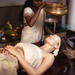 A therapist performing Shirodhara, an Ayurvedic treatment, pouring warm oil from a brass vessel onto the forehead of a woman lying on a wooden massage table.