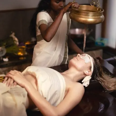 A therapist performing Shirodhara, an Ayurvedic treatment, pouring warm oil from a brass vessel onto the forehead of a woman lying on a wooden massage table.