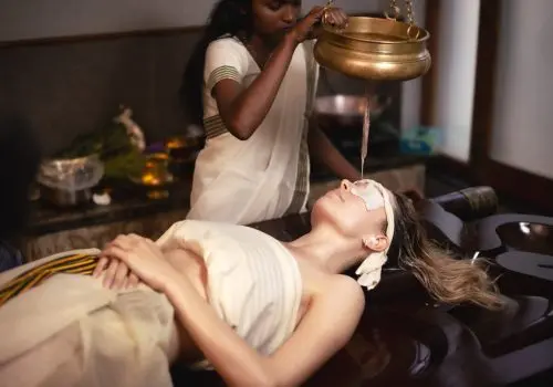 A therapist performing Shirodhara, an Ayurvedic treatment, pouring warm oil from a brass vessel onto the forehead of a woman lying on a wooden massage table.