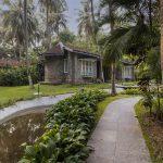 Tropical resort pathway with stone cottages, palm trees, and a small water channel.