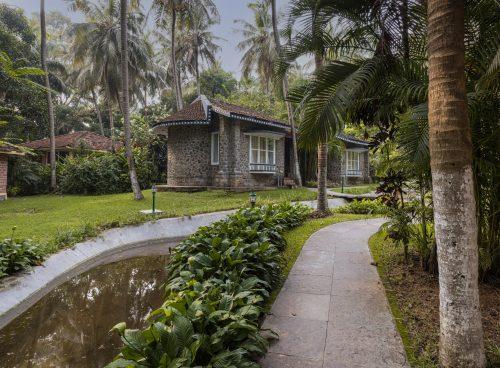 Tropical resort pathway with stone cottages, palm trees, and a small water channel.