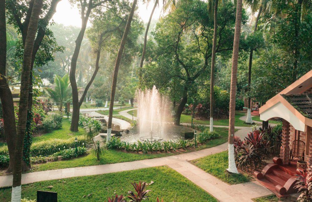 A serene garden at Kairali – The Ayurvedic Healing Village, featuring a central fountain surrounded by lush tropical greenery, winding pathways and traditional Kerala villa.