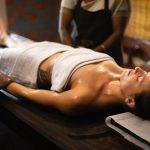 A woman lying on a wooden massage table receiving an Ayurvedic oil massage from two therapists.