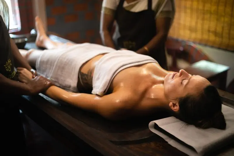 A woman lying on a wooden massage table receiving an Ayurvedic oil massage from two therapists.