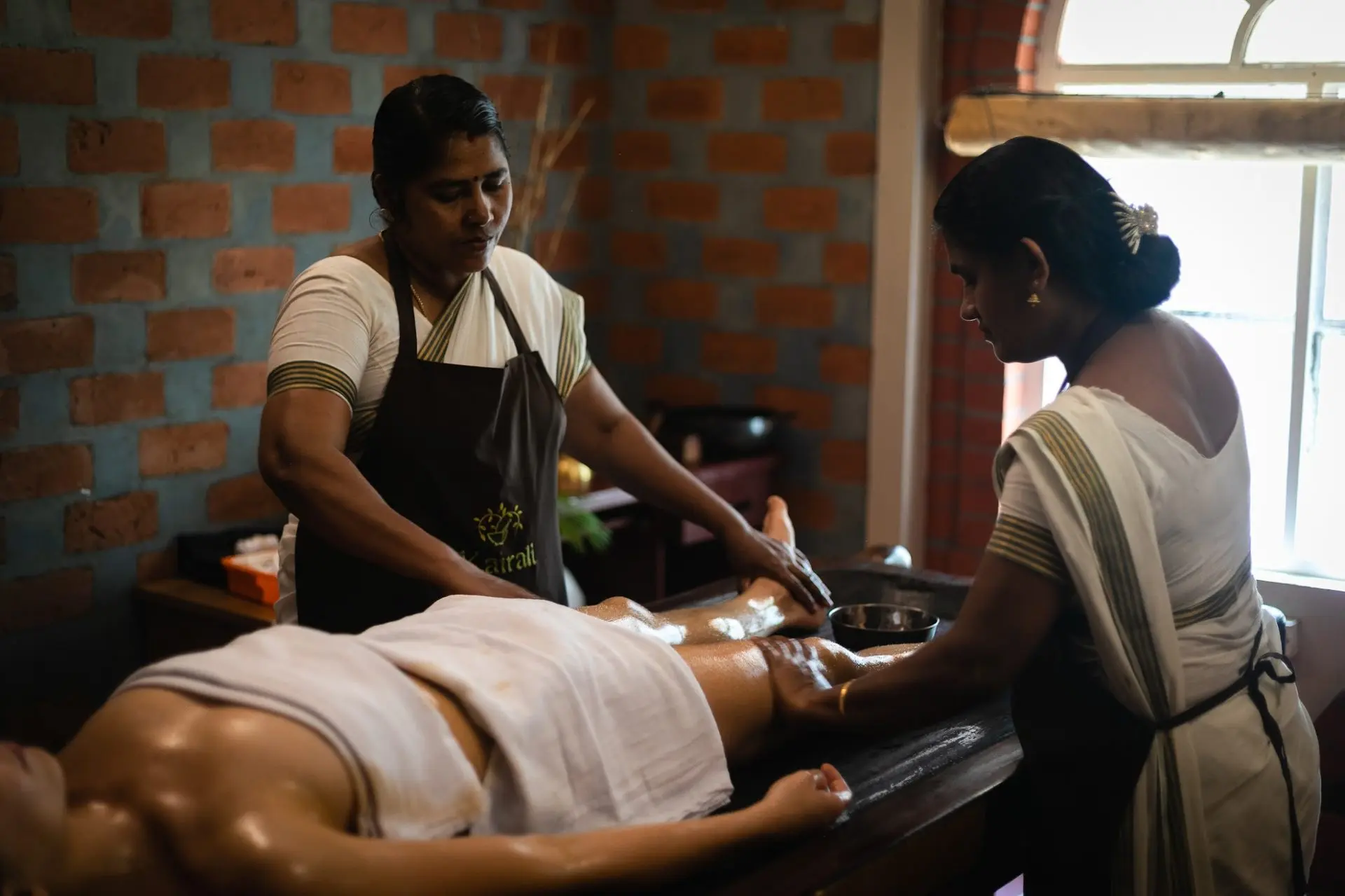 A woman receiving a traditional Ayurvedic massage from two therapists at Kairali spa, lying on a wooden massage table