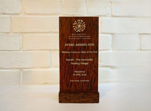 Wooden award plaque from the Asia Pacific Spa & Wellness Coalition, engraved with "APSWC Awards 2026 – Wellness Centre or Clinic of the Year – Kairali – The Ayurvedic Healing Village – Presented 24 April 2026, Siem Reap, Cambodia.