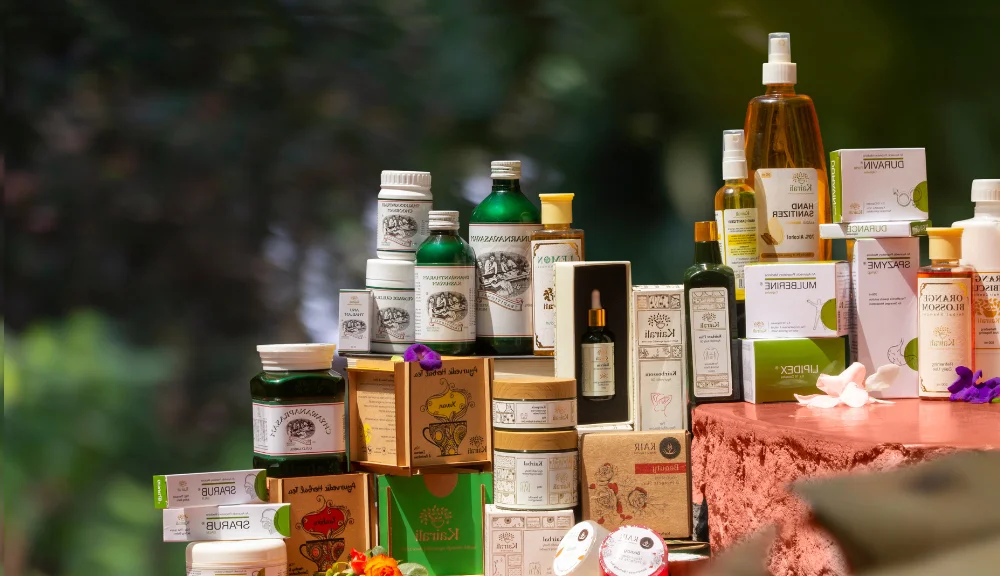 A premium display of Kairali Ayurvedic products including herbal oils, skincare, wellness supplements, and Ayurvedic medicine arranged elegantly with natural floral accents against a green nature-inspired background.