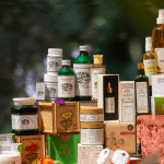 A premium display of Kairali Ayurvedic products including herbal oils, skincare, wellness supplements, and Ayurvedic medicine arranged elegantly with natural floral accents against a green nature-inspired background.