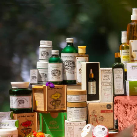 A premium display of Kairali Ayurvedic products including herbal oils, skincare, wellness supplements, and Ayurvedic medicine arranged elegantly with natural floral accents against a green nature-inspired background.