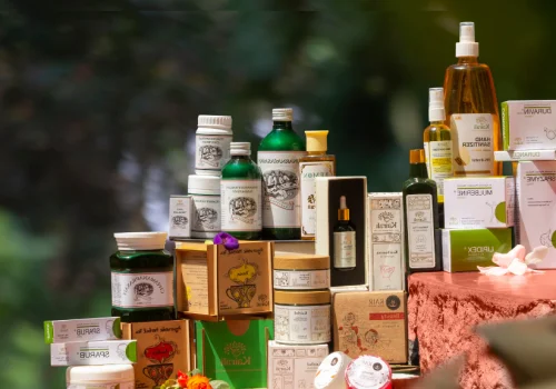 A premium display of Kairali Ayurvedic products including herbal oils, skincare, wellness supplements, and Ayurvedic medicine arranged elegantly with natural floral accents against a green nature-inspired background.