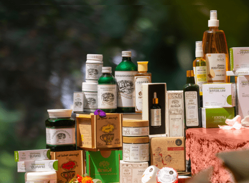 A premium display of Kairali Ayurvedic products including herbal oils, skincare, wellness supplements, and Ayurvedic medicine arranged elegantly with natural floral accents against a green nature-inspired background.
